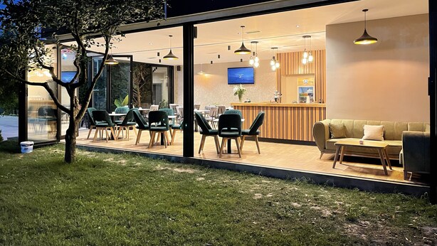 A modern outdoor dining area with glass walls, a wooden counter, and a sofa. - Airport Garden Hotel (Nikël)