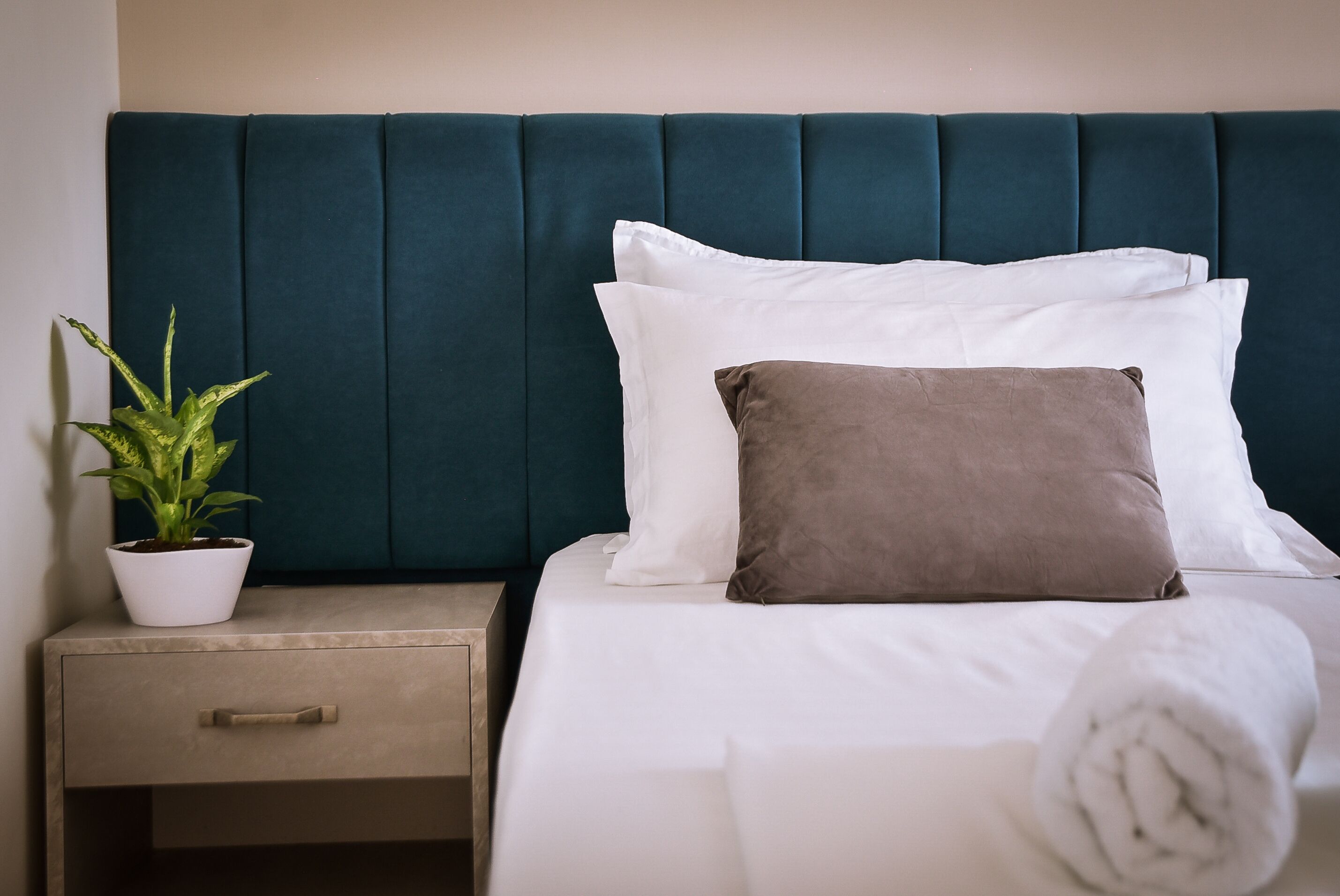 Una cama bien hecha con almohadas blancas y marrones, un cabecero color turquesa y una pequeña maceta sobre una mesita de noche.