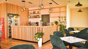 A modern cafe with a wooden counter, green upholstered chairs, and a variety of plants. - Airport Garden Hotel (Nikël)