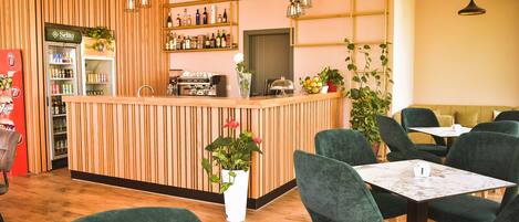 Un café moderne avec un comptoir en bois, des chaises à l’assise rembourrée de couleur verte et une variété de plantes.