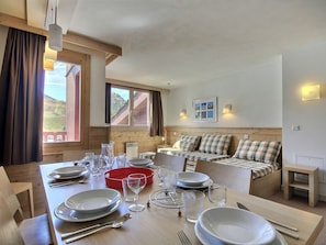 Un comedor con mesa de madera preparada para cuatro personas, un sofá con cojines escoceses, y una ventana con vistas a una montaña.