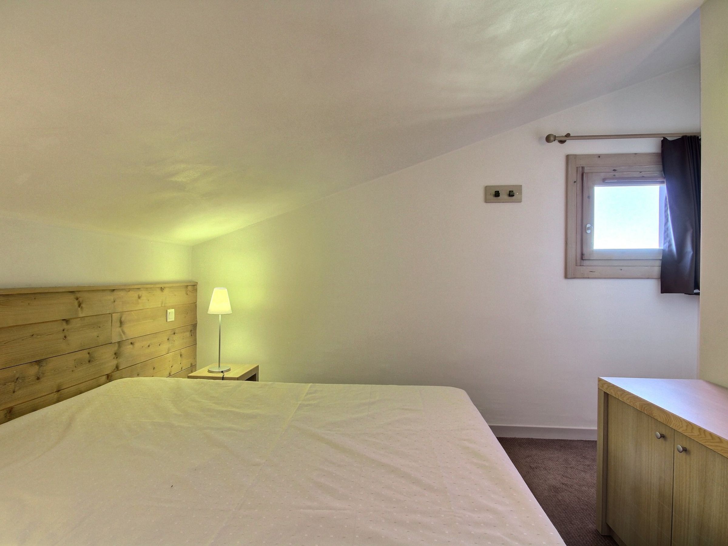 Ein Schlafzimmer mit einem hölzernen Kopfteil, einem Einzelbett, einer Nachttischlampe, einem Fenster mit Vorhang und einem kleinen Schrank.