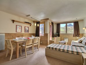 Una habitación acogedora con una mesa de comedor de madera, sillas, una cama con ropa de cama escocesa, un televisor y una ventana con vistas a un paisaje montañoso.