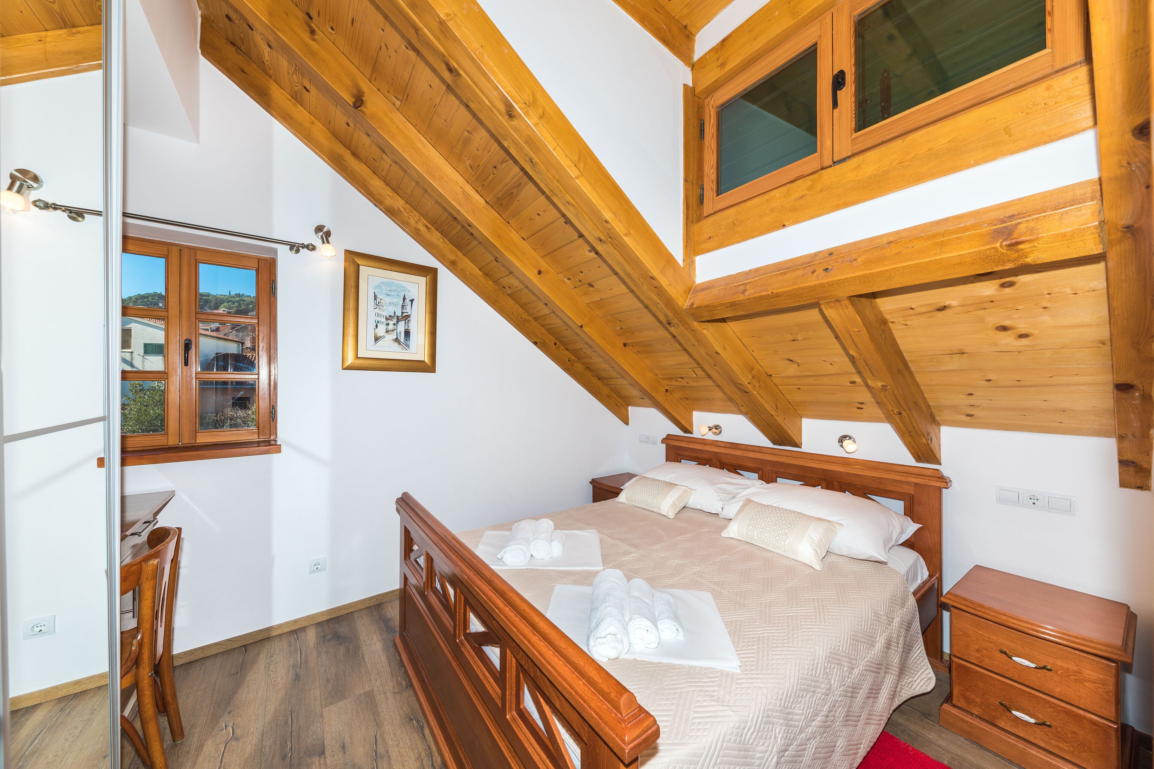 A bedroom with a wooden ceiling, a bed with white linens, a wooden nightstand, and a window with a view of buildings.