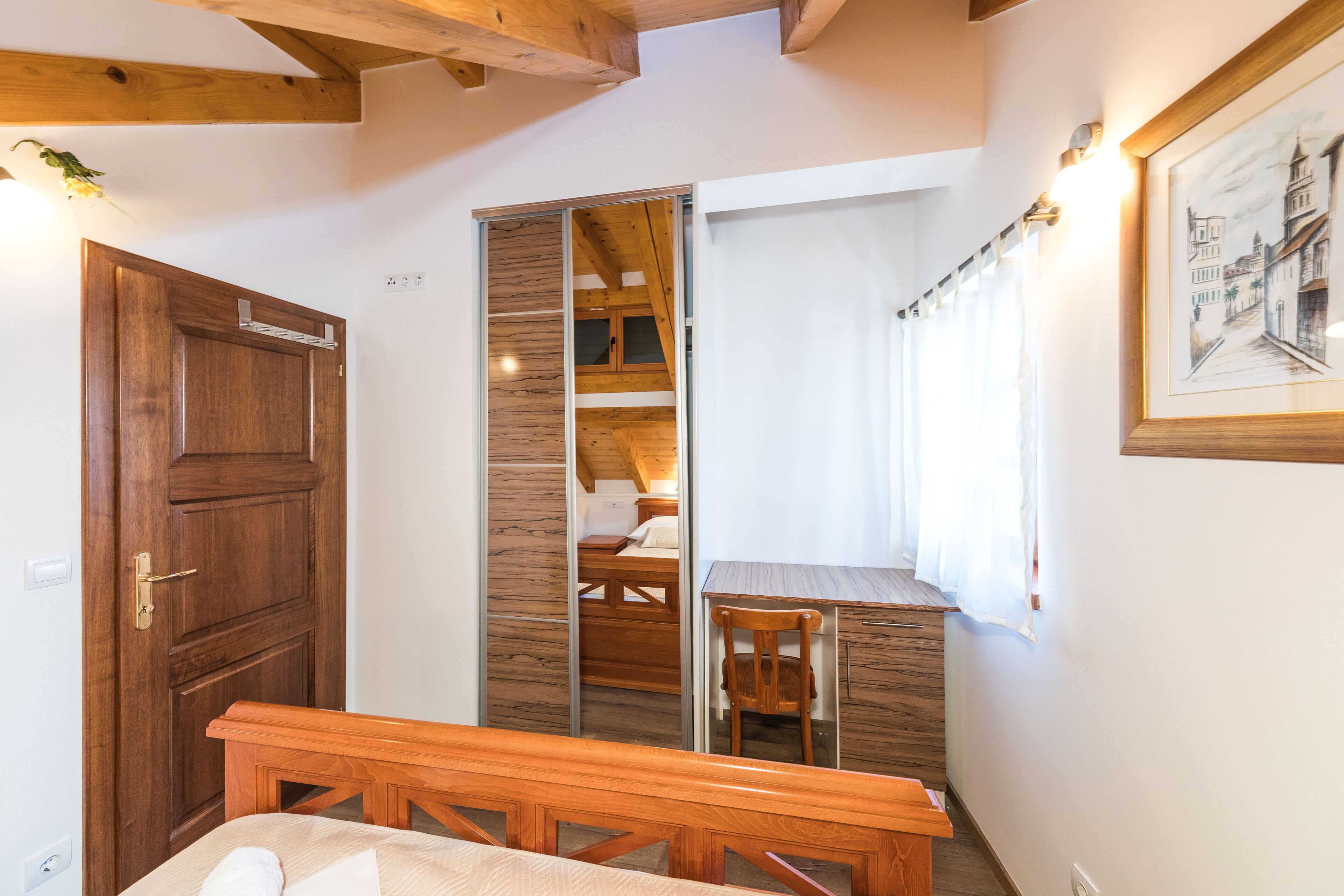 A bedroom with a wooden bed, a desk, and a chair. There is a wardrobe with a mirror and a window with curtains.