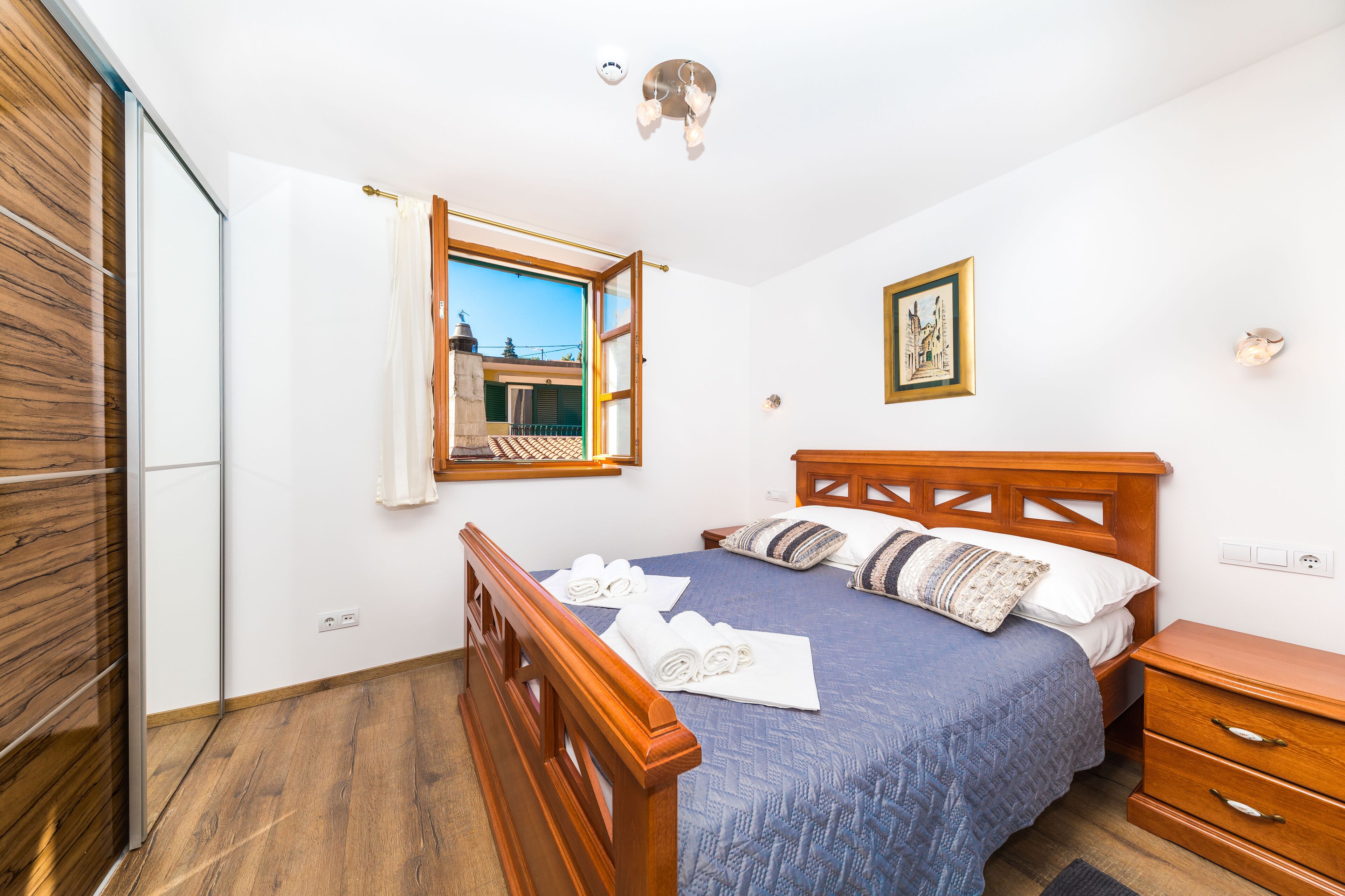 A bedroom with a wooden bed, a window with a view, a bedside table, and a wall-mounted light fixture.