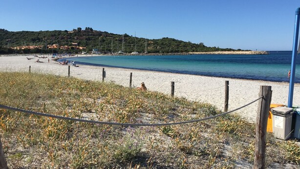 Ein Strand mit sandigem Ufer, blauem Meer und hügeliger Kulisse.