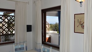 Ein Zimmer mit Tisch und Stühlen, einem fenster mit Vorhängen, Blick ins Freie und einer Wandleuchte.