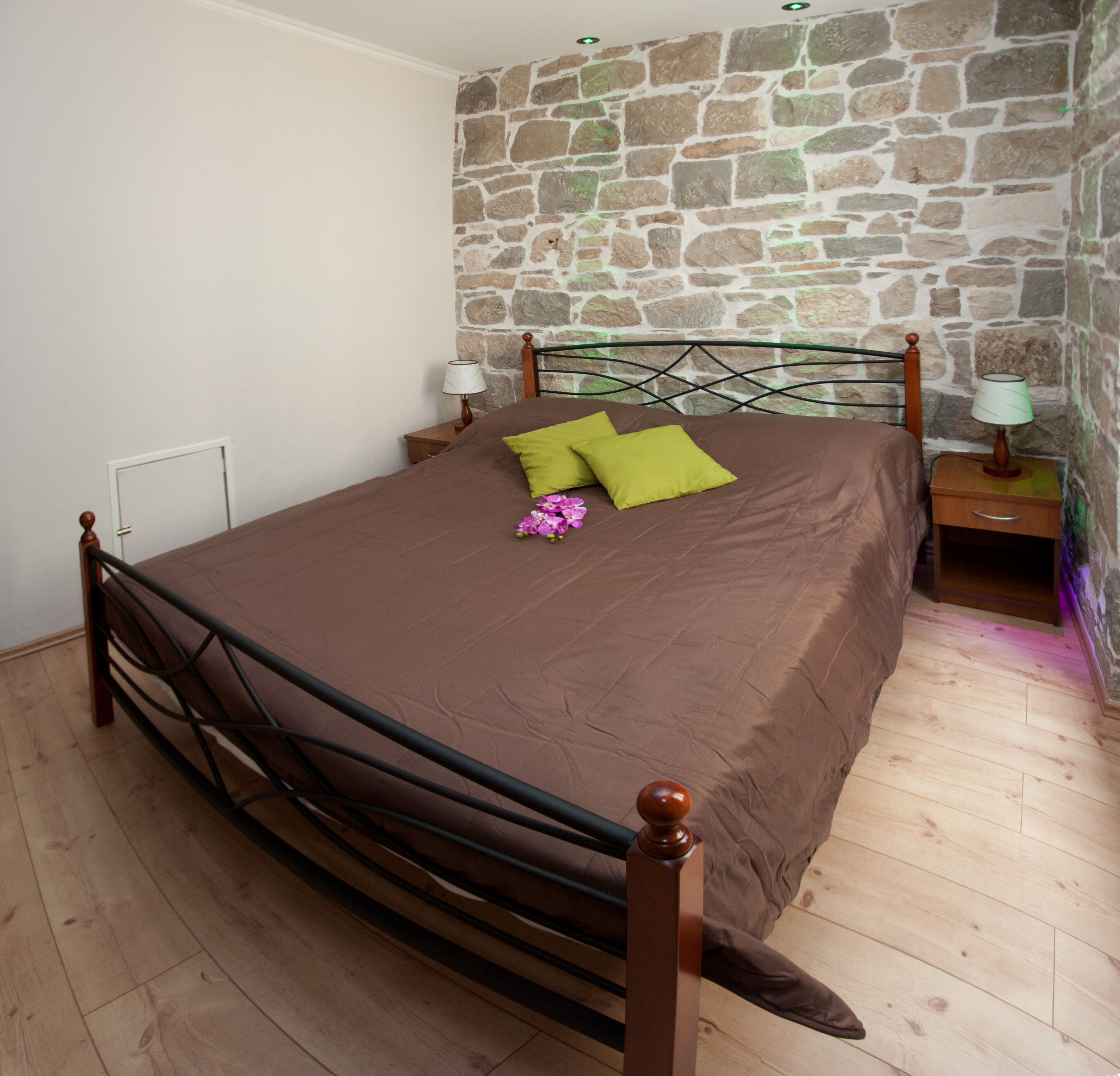 A bedroom with a stone wall, a wooden bed frame, a brown bedspread, two green pillows, a nightstand with a lamp, and a wooden floor.