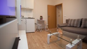 Ein modernes Wohnzimmer mit einem Glastisch, einer Couch mit Karokissen und einer Küchenzeile mit weißen Schränken sowie einem kleinen Esstisch.