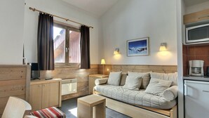 En kompakt opholdsstue med sofa, lille bord, tekøkken og et vindue med gardiner.