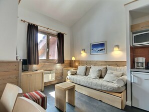 Ein kompakter Wohnbereich mit einer Couch, einem kleinen Tisch, einer Küchenzeile und einem Fenster mit Vorhängen.