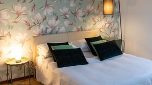 Een net opgemaakt bed met witte lakens en groene en zwarte kussens, een wand met bloemenbehang en een bedlampje.
