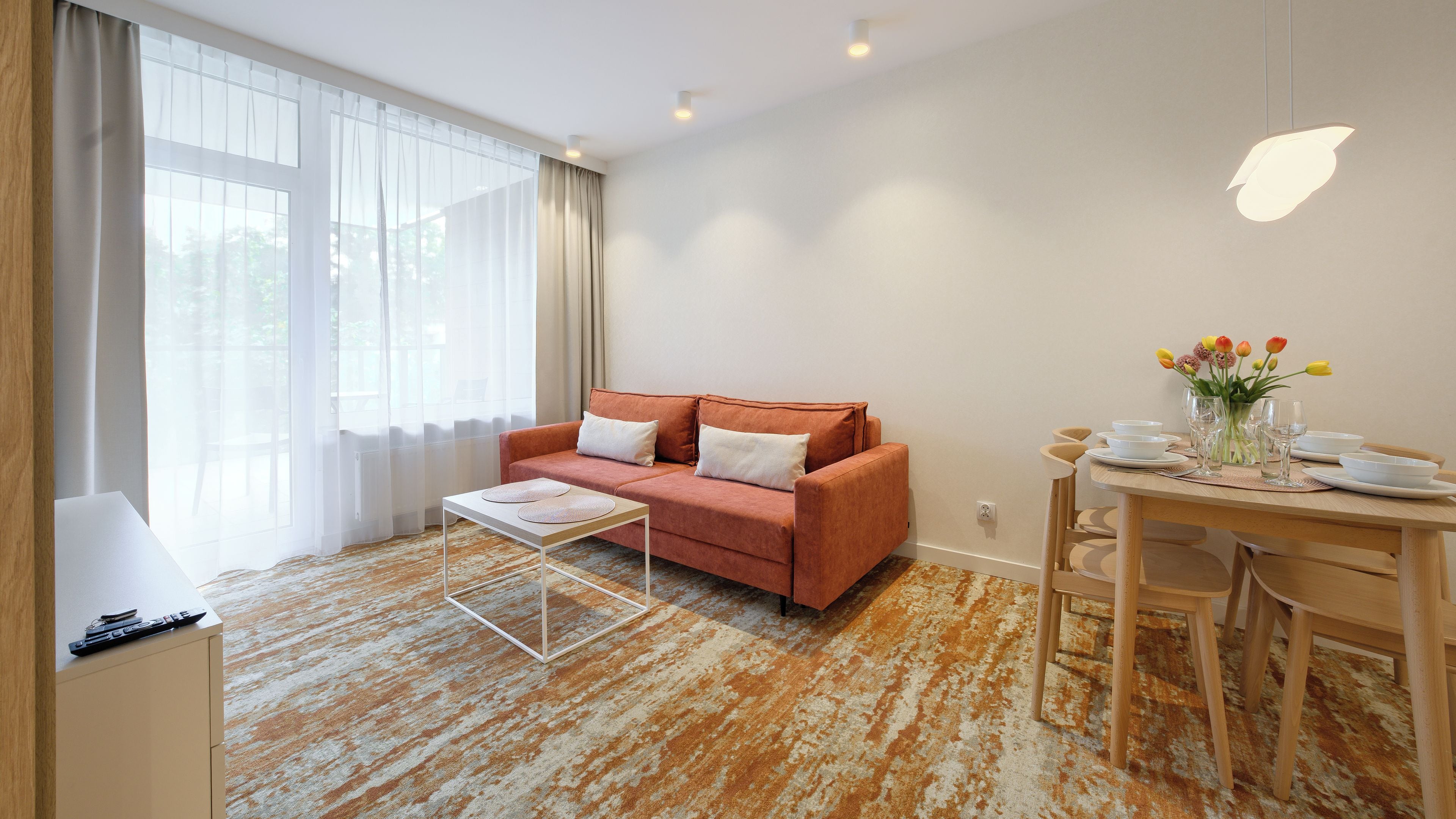 Een moderne woonkamer met een bank, een eettafel voor twee personen en een groot raam met dunne gordijnen.