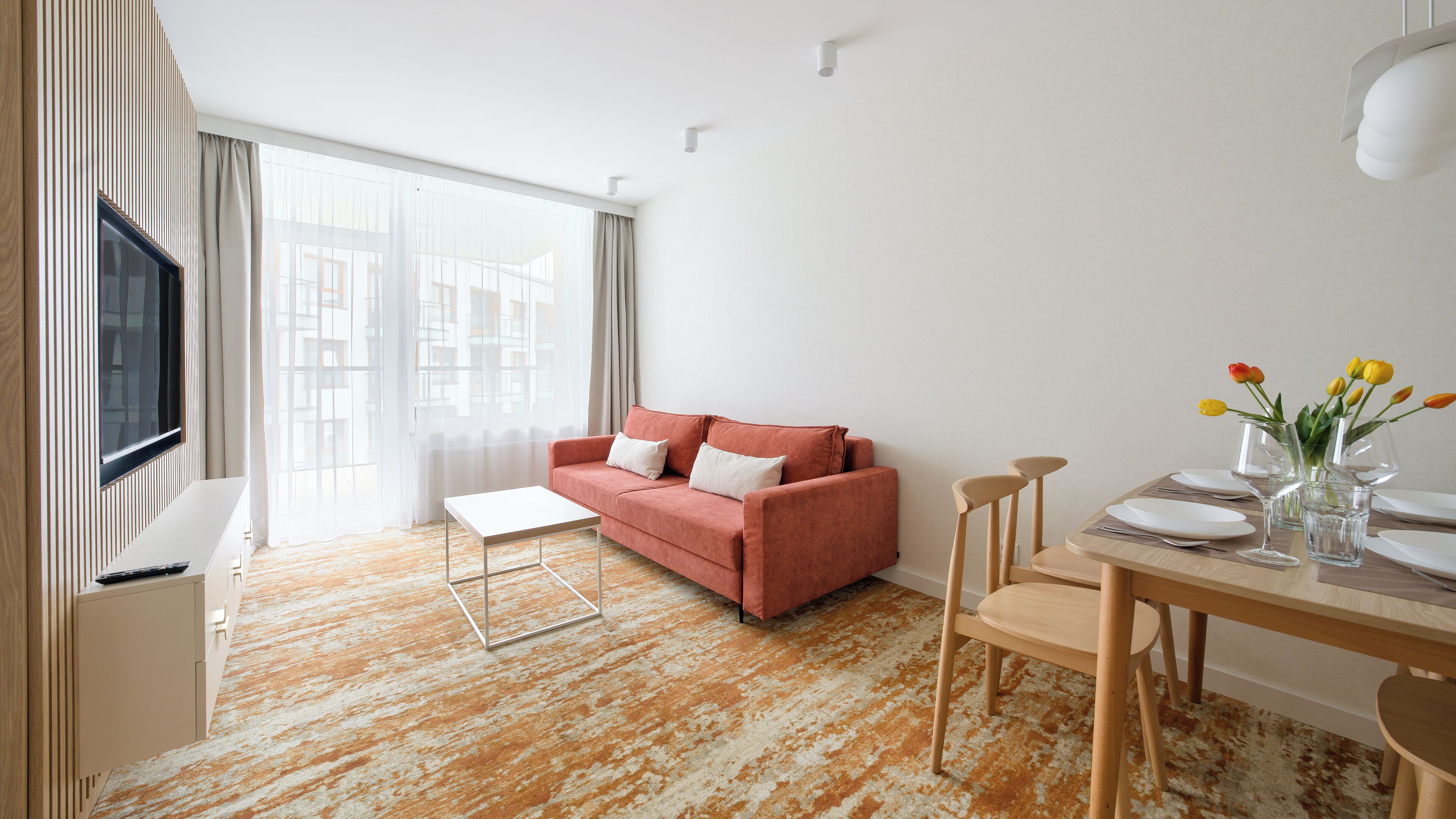 Un salon moderne comprenant un canapé, une table basse, une table à manger avec des chaises, un téléviseur fixé au mur et une fenêtre avec des rideaux.