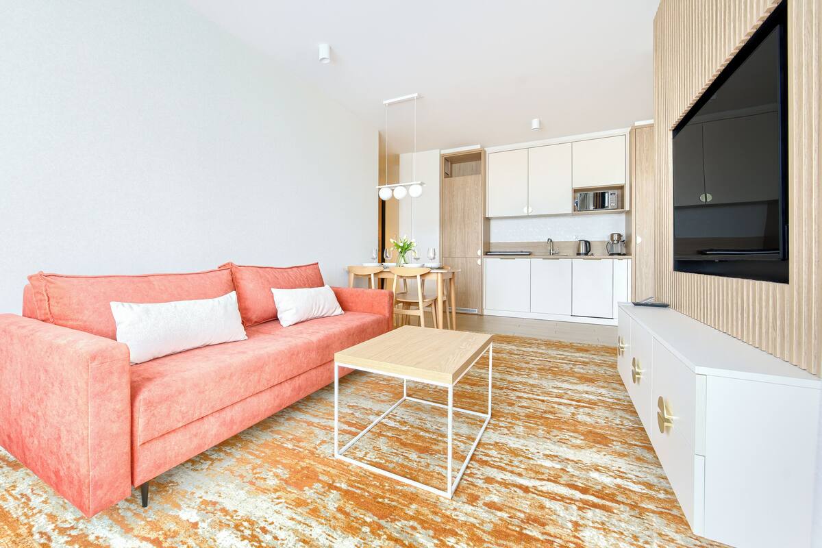 A modern living room with a pink sofa, a wooden coffee table, and a kitchen area with white cabinets and a TV.
