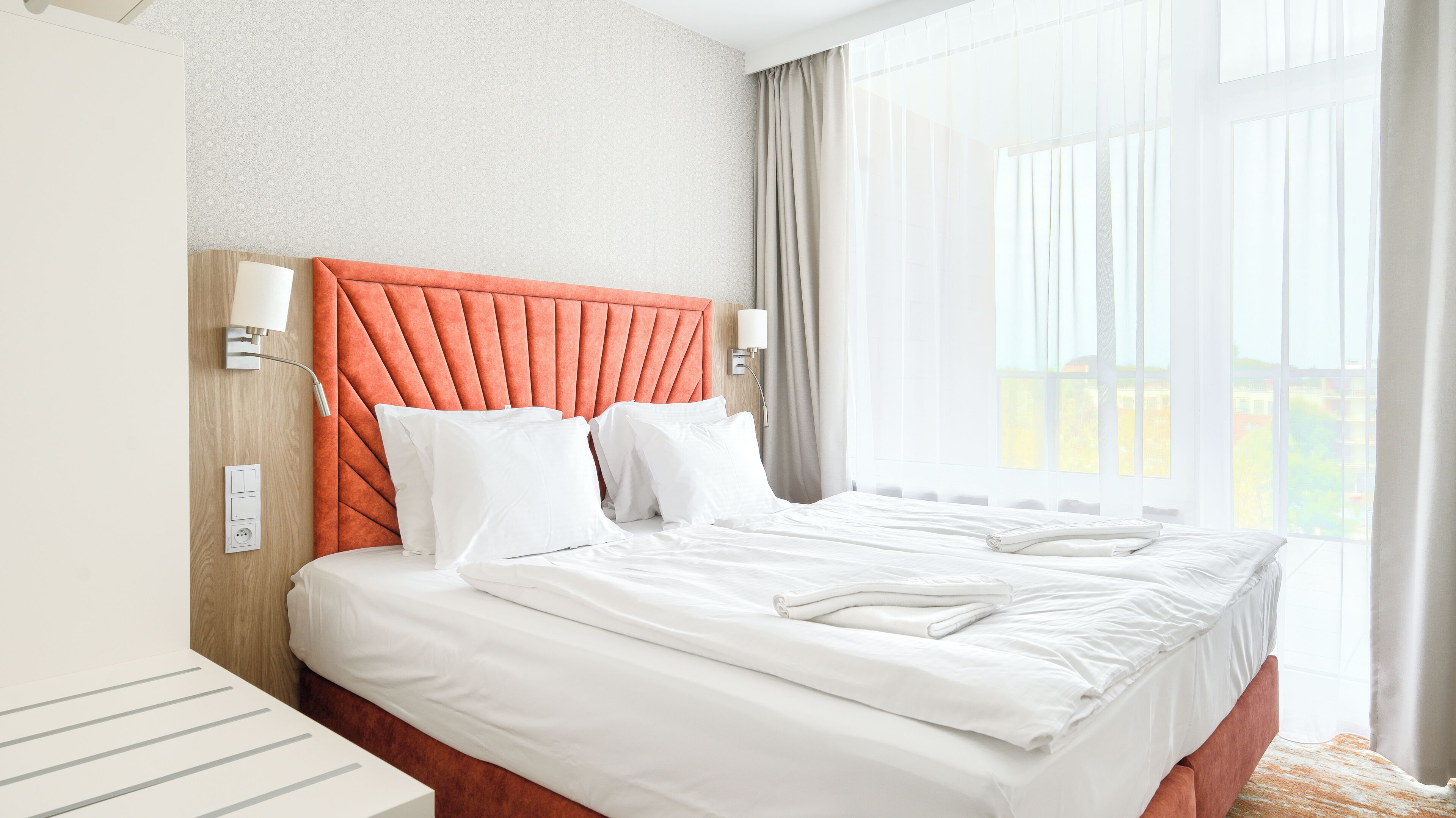 A hotel room with a bed, a headboard, a wall-mounted lamp, and a window with curtains.