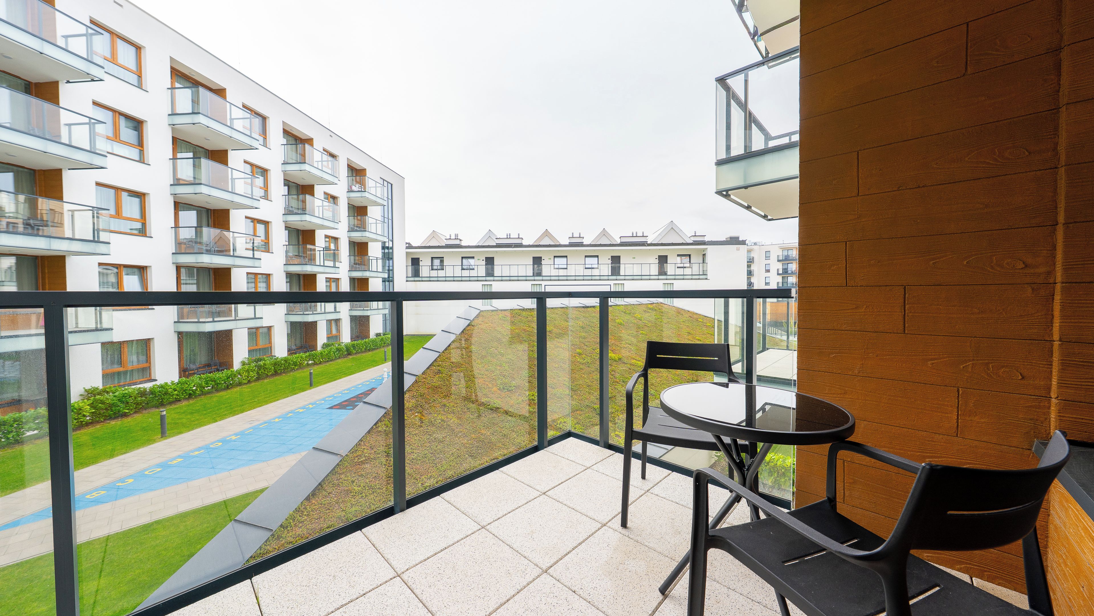 Un balcon doté d’une balustrade en verre, d’une table et de deux chaises, offrant une vue sur une piscine et un espace verdoyant, avec des bâtiments en arrière-plan.