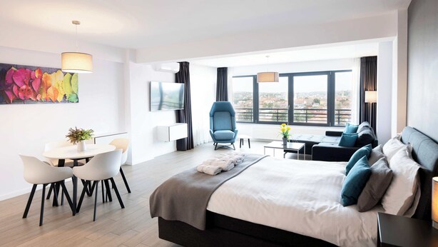 A modern hotel room with a large bed, a seating area with a blue chair, a dining table with chairs, and a city view through large windows.