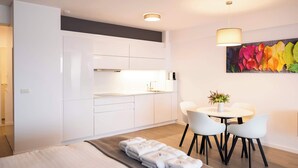 A modern hotel room with a bed, a dining area with a table and chairs, a kitchenette, and a colorful flower painting on the wall.