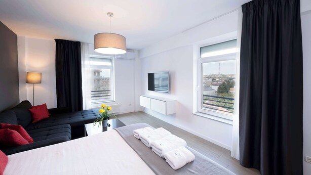 A modern hotel room with a bed, a sofa, a TV, and large windows offering a city view.