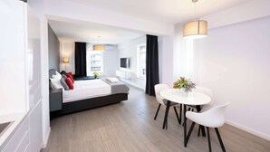 A modern hotel room with a bed, bedside table, lamp, and a dining area with a table and chairs.