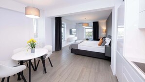 A modern hotel room with a bed, a dining area with a table and chairs, and a large window providing natural light.
