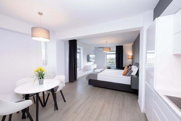 Quarto de hotel moderno com cama, área de refeições com mesa e cadeiras, e uma janela grande que proporciona luz natural.