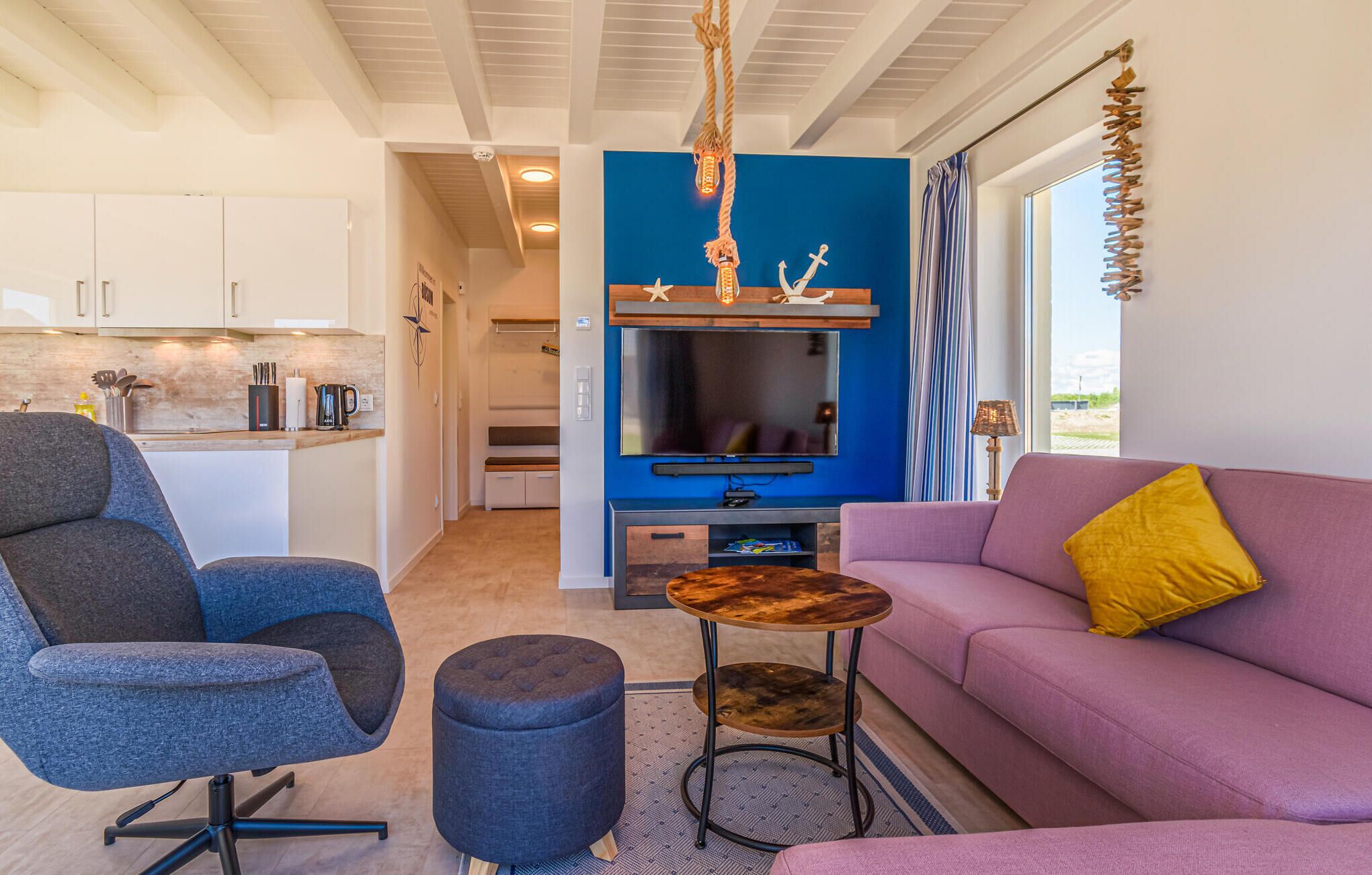 Ein modernes Wohnzimmer mit einer Couch, einem Sessel, einem Couchtisch und einem Fernseher. Im Hintergrund ist eine Küchenzeile mit Schränken und einer Arbeitsplatte zu sehen.
