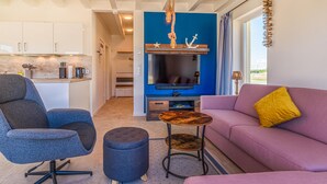 Ein modernes Wohnzimmer mit einer Couch, einem Sessel, einem Couchtisch und einem Fernseher. Im Hintergrund ist eine Küchenzeile mit Schränken und einer Arbeitsplatte zu sehen.