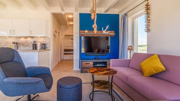 Ein modernes Wohnzimmer mit einer Couch, einem Sessel, einem Couchtisch und einem Fernseher. Im Hintergrund ist eine Küchenzeile mit Schränken und einer Arbeitsplatte zu sehen.