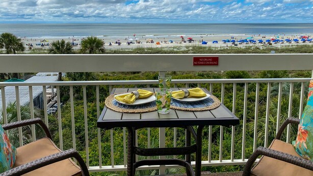 A balcony with a table set for two, overlooking a beach with umbrellas and people. A sign on the railing reads "NO GRILLING NO TOWELS ON RAILING."