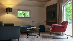 Ein modernes Wohnzimmer mit einer Couch, einem roten Sessel, einem kleinen runden Tisch, einer Stehlampe, einem Fenster mit Blick auf Grünflächen und einem an der Wand befestigten Fernseher.