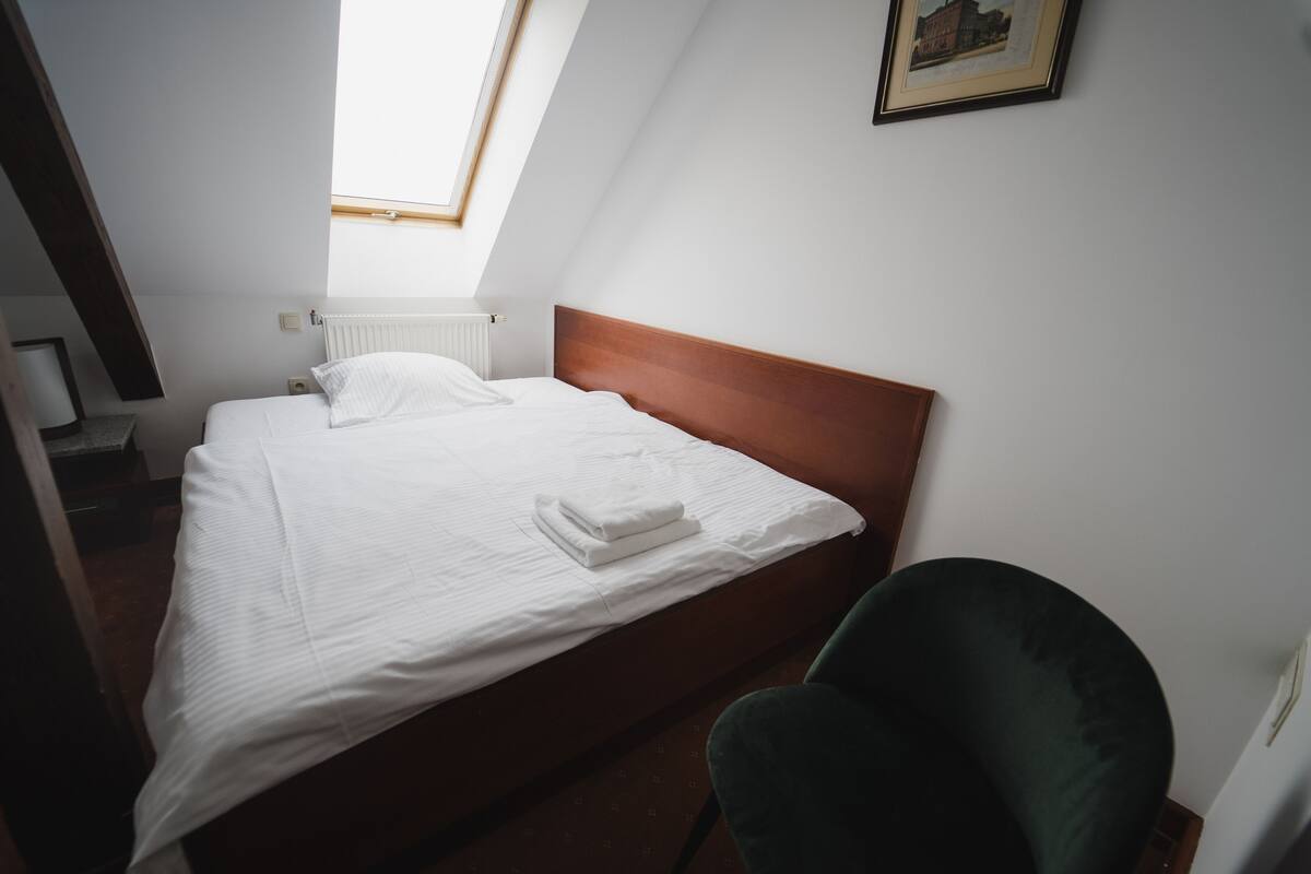 Una cama bien hecha con sábanas blancas, cabecera de madera y una silla verde en una habitación con techo inclinado y ventana.