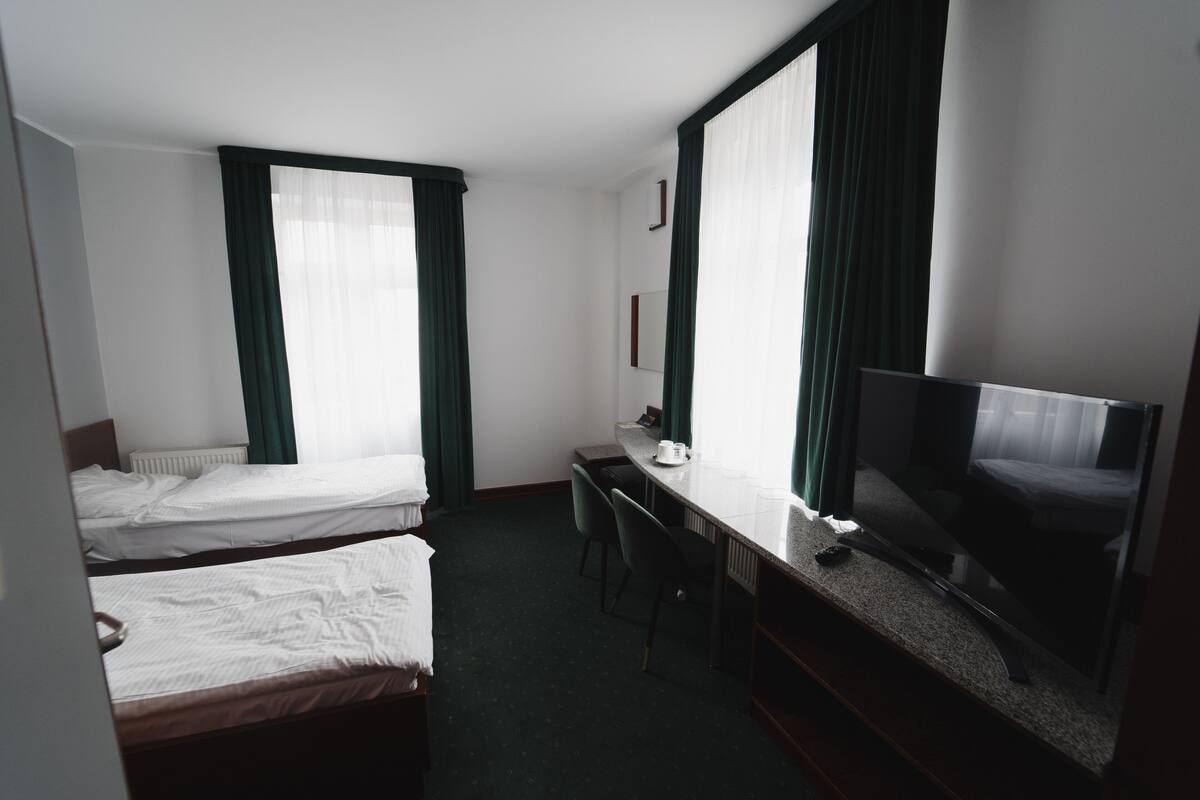 Habitación de hotel con dos camas individuales, un escritorio, una silla, un televisor y un ventanal grande con cortinas.