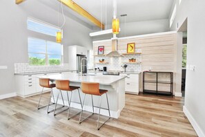 A modern kitchen with a large island, bar stools, pendant lights, and a view of the outdoors. - River Road Retreat - NEW Modern 3 Bedroom on the Blanco River (Wimberley)