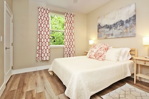 A bedroom with a bed, two bedside tables, a window with patterned curtains, a painting on the wall, and a wooden floor. - River Road Retreat - NEW Modern 3 Bedroom on the Blanco River (Wimberley)