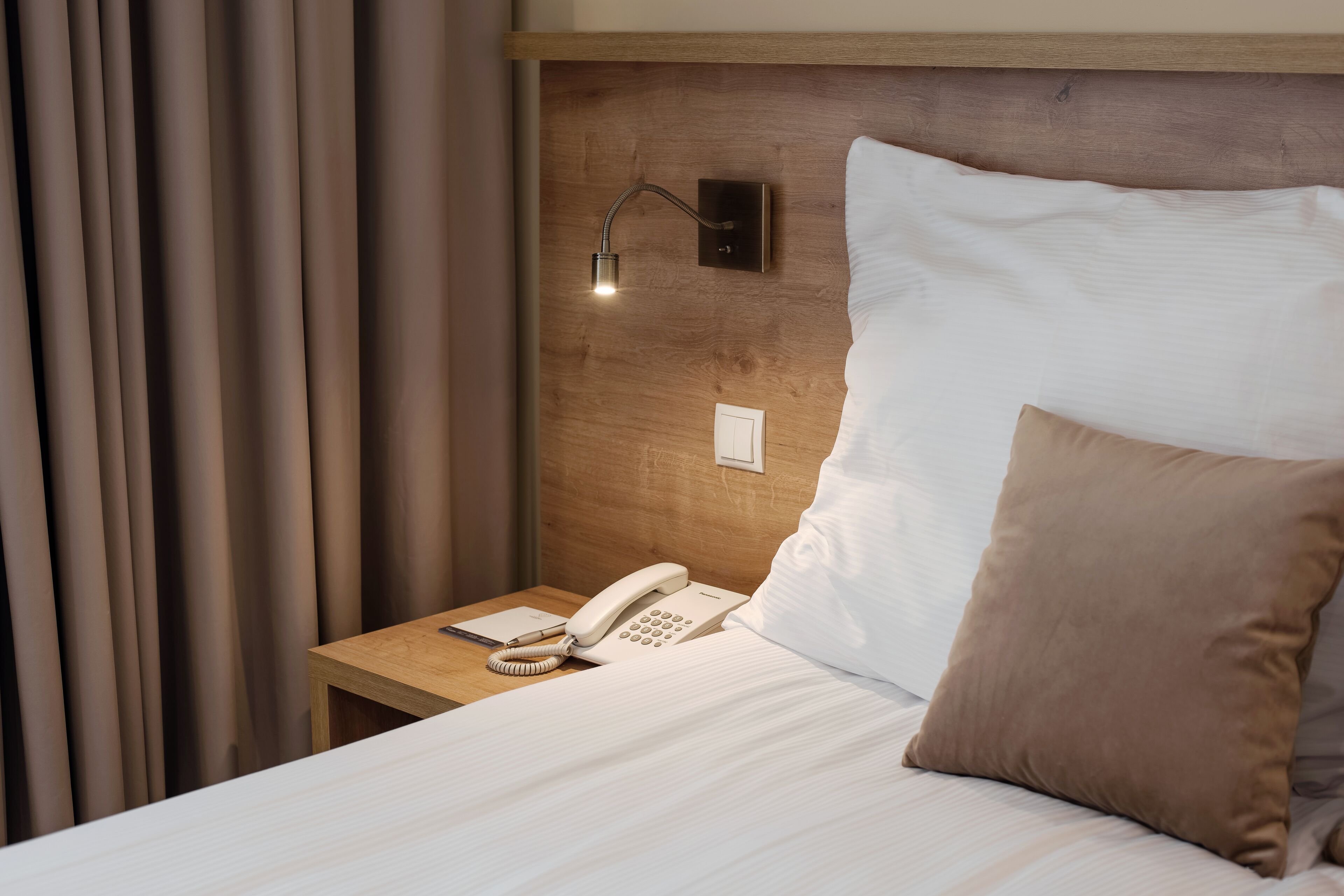 A neatly made bed with a white comforter and a brown pillow, a bedside table with a white telephone, and a wall-mounted light fixture.