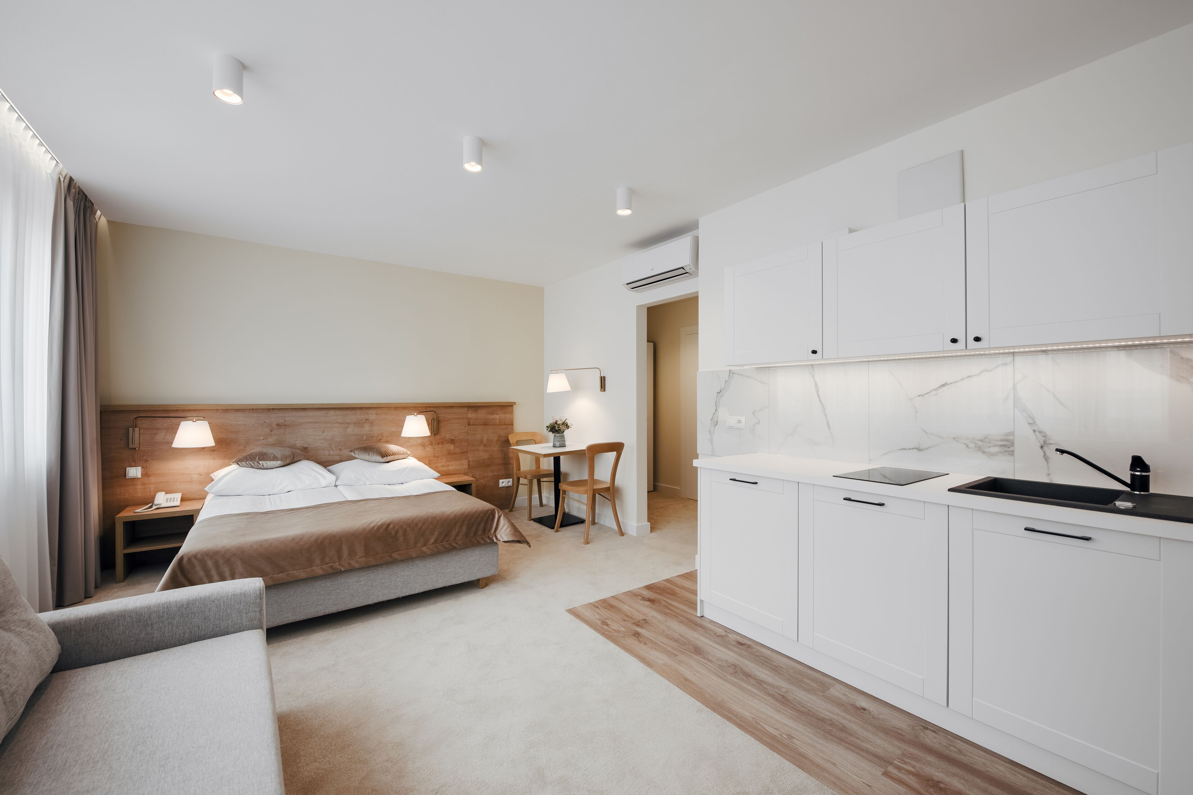 Habitación de hotel moderna con cama, mesita de noche, lámpara y una kitchenette con lavabo y armarios.