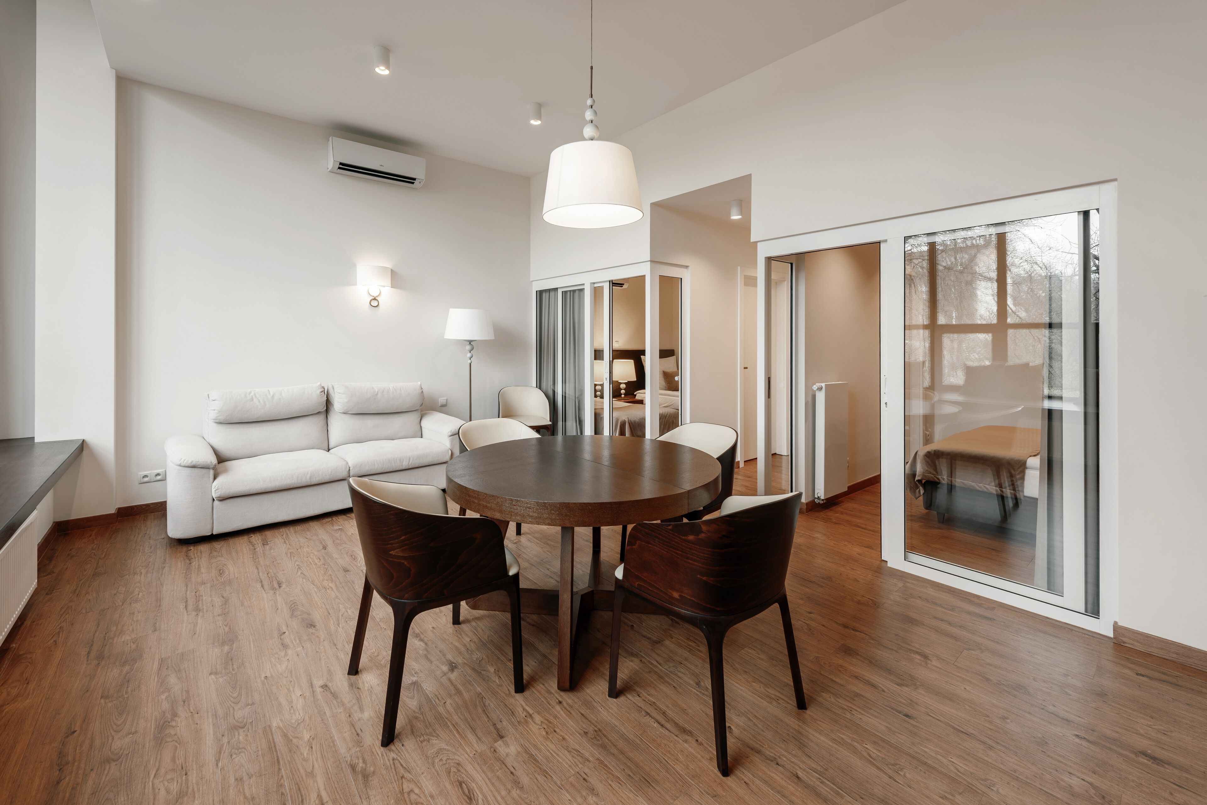 A modern living room with a round wooden table, dark chairs, a white sofa, and a large window.
