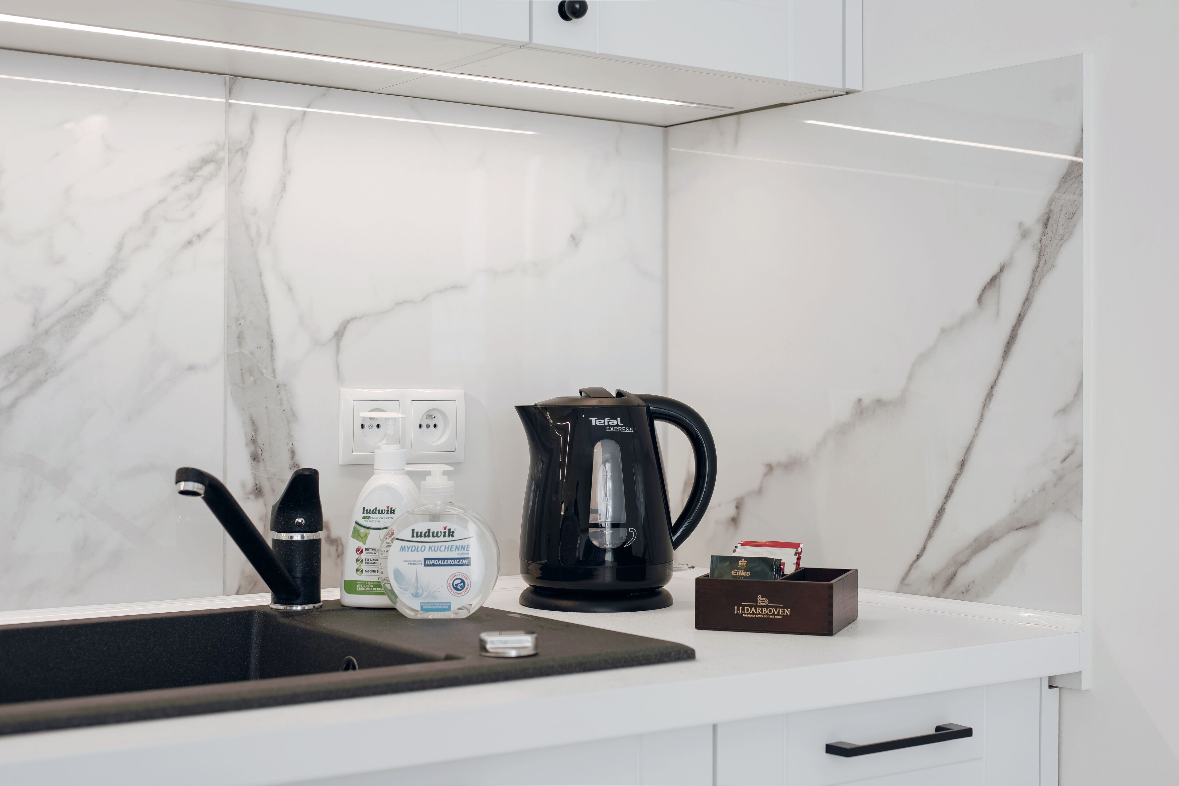 A modern kitchen with a marble backsplash, a black coffee maker, a bottle of hand soap, and a box of coffee pods.