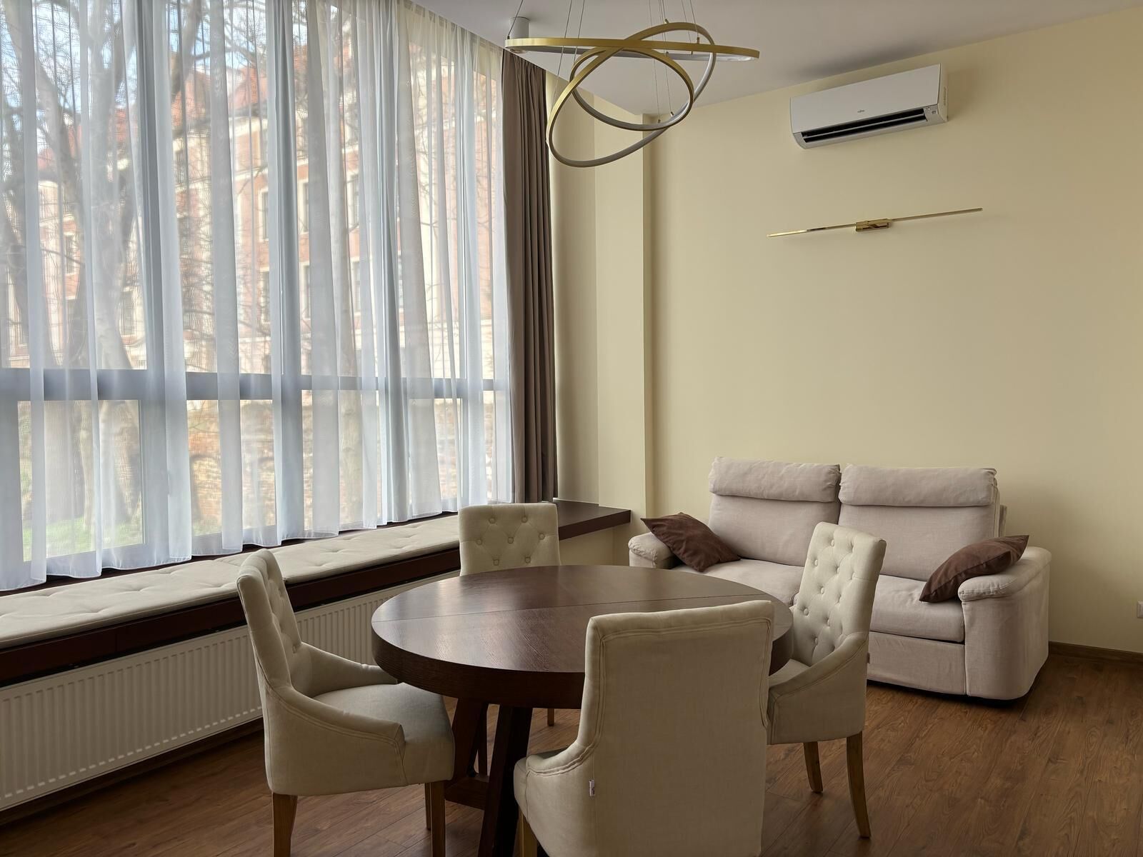 A modern living room with a round wooden table, four white chairs, a beige sofa with two cushions, and a large window with sheer curtains.