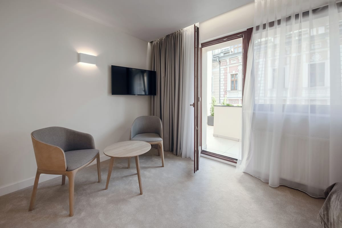 Een moderne kamer met een flatscreen televisie aan de muur, twee stoelen, een klein rond tafeltje en een groot raam met dunne gordijnen.
