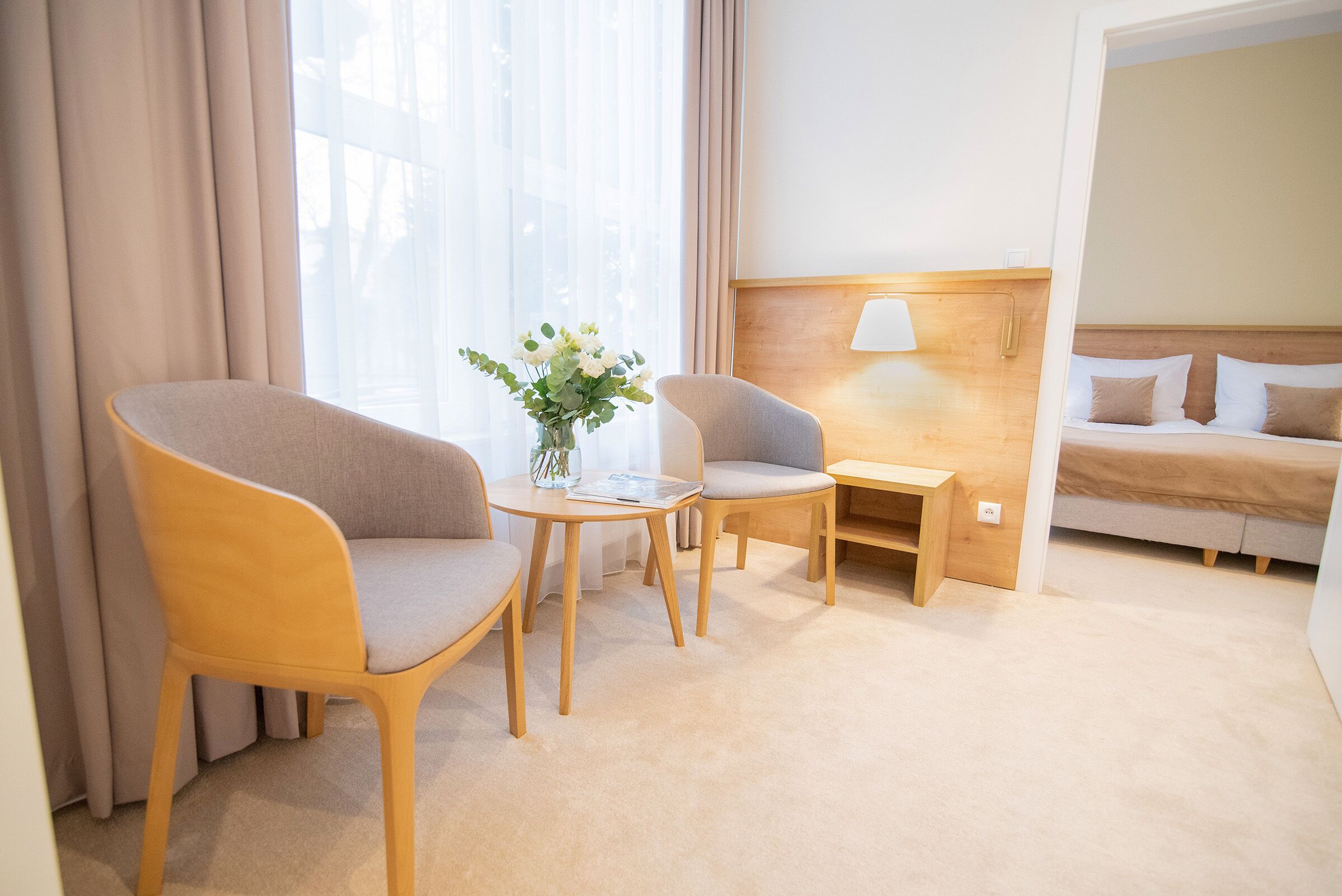 Ein Hotelzimmer mit Bett, zwei Sesseln, einem kleinen Tisch mit Blumenvase, einem Nachttisch mit Lampe und Blick auf die Stadt durch große Fenster.