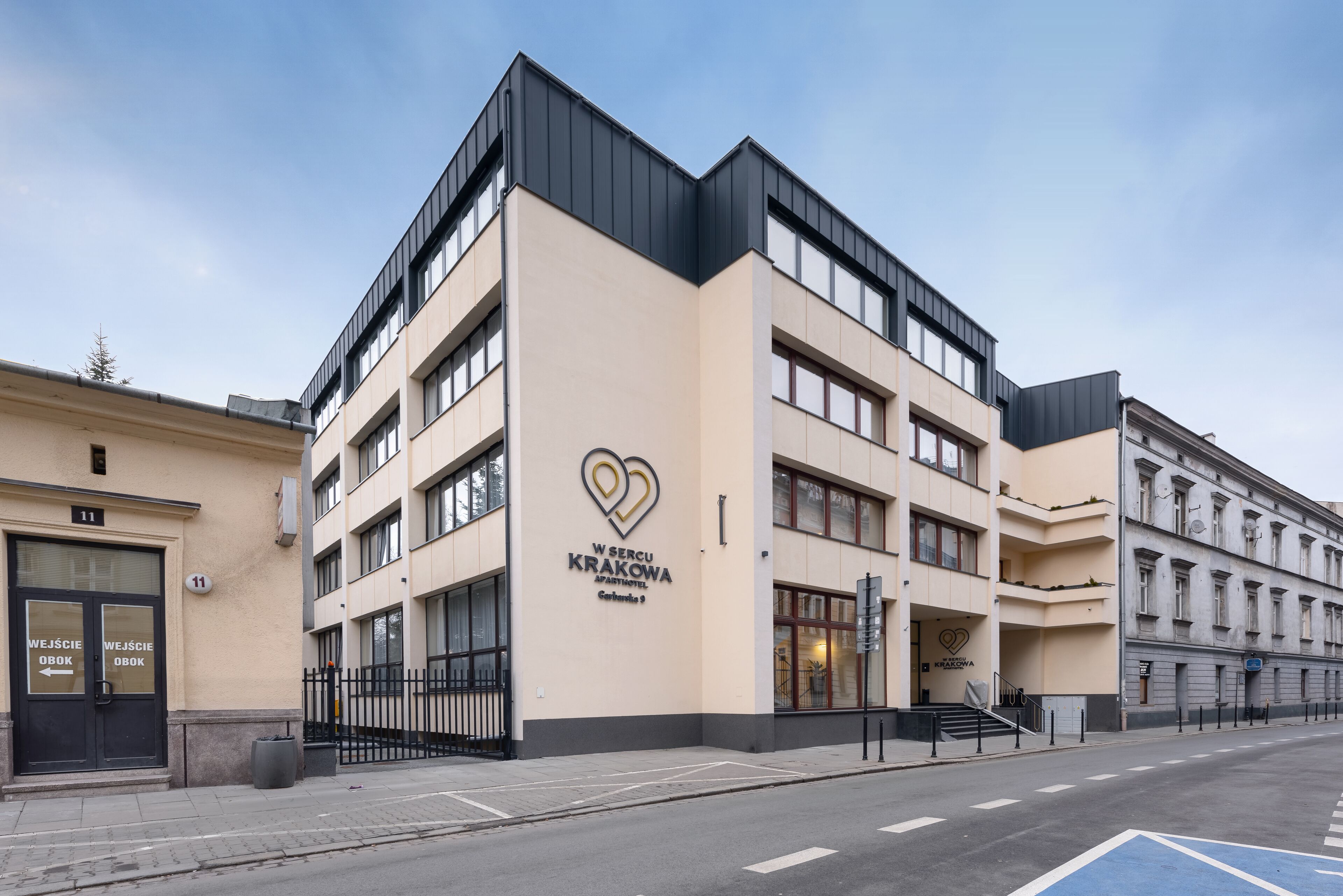A modern building with a heart-shaped logo, labeled "W SERCU KRAKOWA APARTMENTS," featuring multiple floors and a prominent entrance.