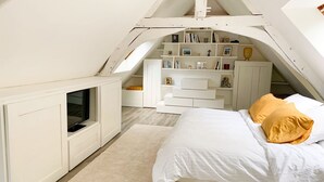 A bedroom with a sloped ceiling, built-in bookshelves, a bed with white bedding and two pillows, and a small seating area with a white cabinet. - House in Perche (Marolles-les-Braults)