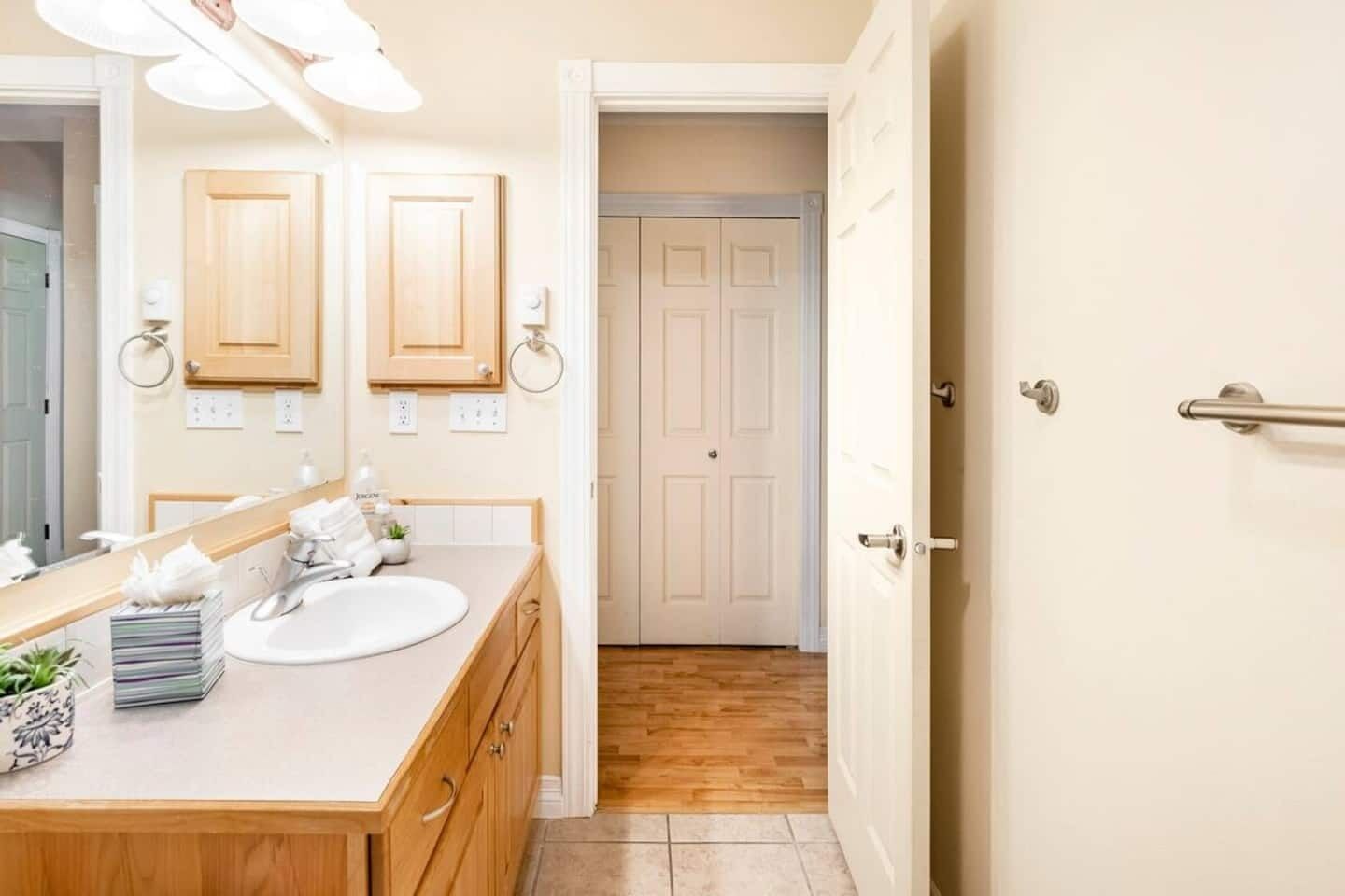 A bathroom with a wooden vanity, a white sink, and a mirror. There is a towel rack above the mirror and a door leading to another room.