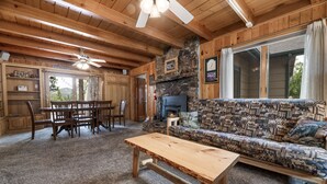 A cozy cabin interior with a stone fireplace, wooden furniture, and a large window with curtains. - Beautiful Back Yard with peekaboo Lake Views. Private Hot Tub & Pool Table (Big Bear Lake)
