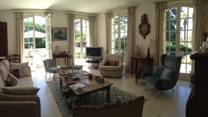 Een ruime woonkamer met een bank, fauteuils, een salontafel en een televisie. Grote ramen zorgen voor daglicht en uitzicht op de omgeving.