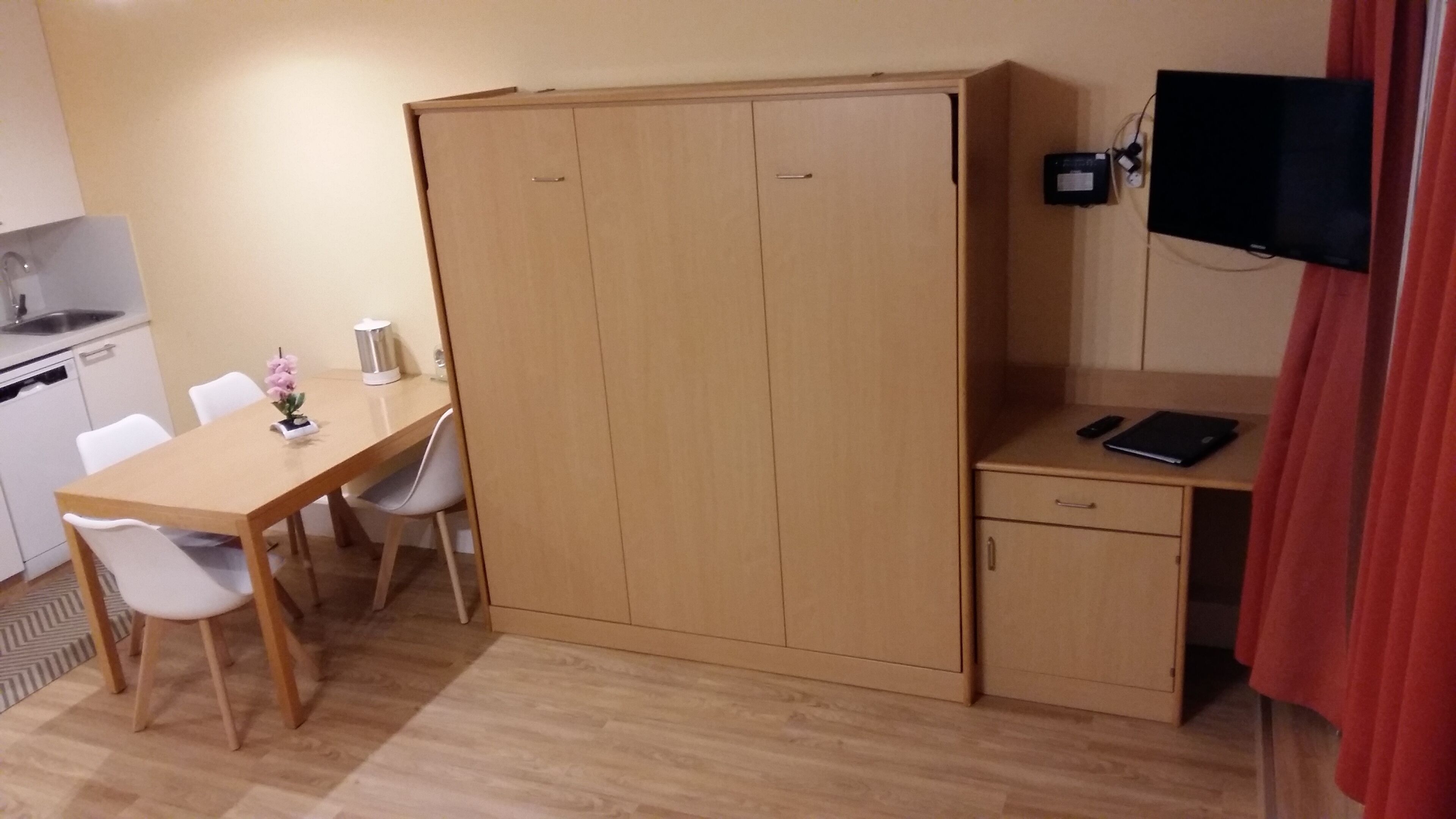 Una habitación compacta con un escritorio de madera, una silla, una pequeña kitchenette, un armario y un televisor montado en la pared.